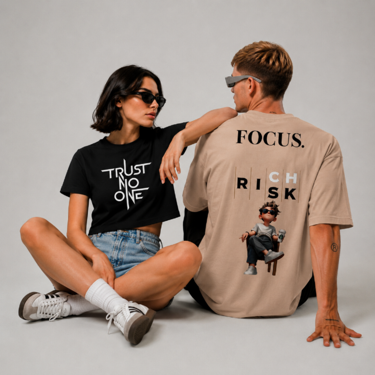 Two models sitting on a light gray studio floor wearing oversized graphic t-shirts. The woman wears a black cropped t-shirt with 'TRUST NO ONE' in white text, while the man wears a beige oversized t-shirt showing the back graphic with 'FOCUS.